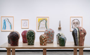 Hand-painted sculptures on table with framed ink drawings in background.